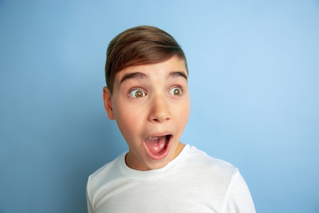 Conmocionado gritando. De cerca. Retrato de niño caucásico aislado sobre fondo azul de estudio. Modelo masculino adolescente hermoso en la presentación de la camisa blanca. Concepto de emociones humanas, expresión facial, ventas, publicidad.