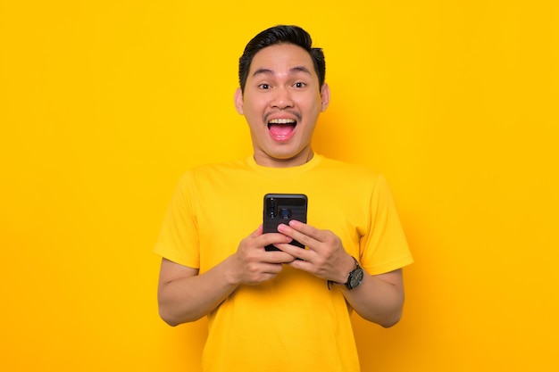 Conmocionado joven asiático en camiseta casual sosteniendo el teléfono móvil y mirando a la cámara aislada sobre fondo amarillo Concepto de estilo de vida de la gente