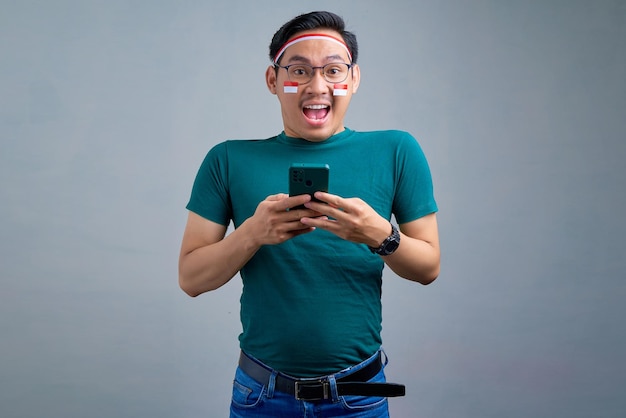 Conmocionado joven asiático en camiseta casual sosteniendo el teléfono móvil y mirando a la cámara aislada sobre fondo gris concepto de celebración del día de la independencia de Indonesia