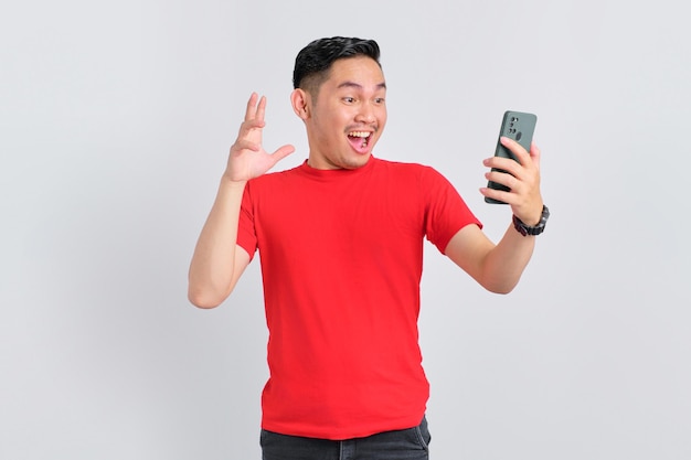 Conmocionado joven asiático mirando la pantalla del teléfono móvil reaccionando a las noticias en línea con la boca abierta aislado sobre fondo blanco.