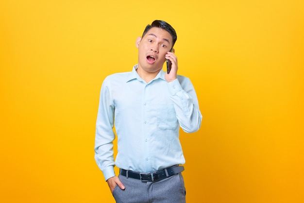 Conmocionado joven empresario guapo hablando por un teléfono inteligente sobre fondo amarillo