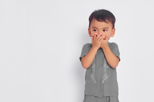 Conmocionado niño musulmán asiático cubriendo la boca con las manos y mirando el espacio vacío aislado en el fondo del estudio blanco