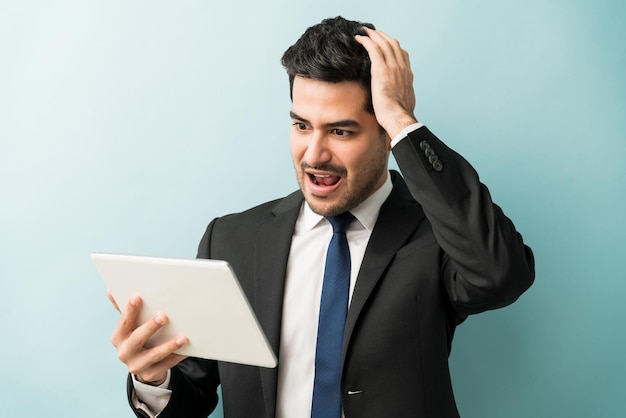 Conmocionado profesional masculino con la mano en la cabeza mientras mira la tableta digital contra el fondo azul.