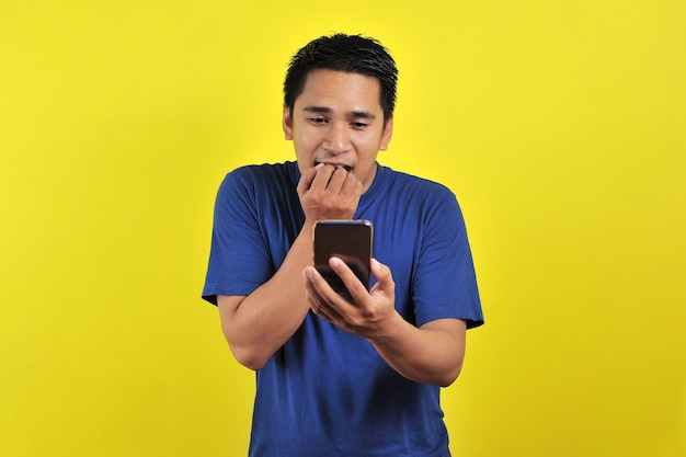 Conmocionado rostro de hombre asiático con camisa blanca mirando la pantalla del teléfono sobre fondo amarillo.