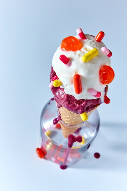 Cono de galleta con helado de vainilla afrutado decorado con diferentes cápsulas en una botella de vidrio sobre un fondo azul con gotas de helado. Vista superior