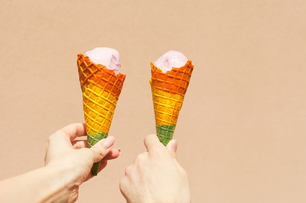 Cono de helado arco iris - dos manos sosteniendo conos de helado en colores del arco iris en un caluroso día de verano cerca de fondo rosa