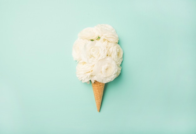 Cono de waffle con flores de ranúnculo blanco sobre fondo azul pastel