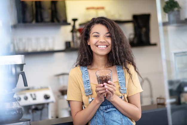 Conocedor del café. Mujer feliz con cabello largo oscuro en ropa casual con porción de granos de café en vidrio en café