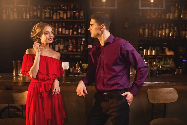 Conocido en el bar, hermosa mujer en vestido rojo coquetea con el hombre detrás del mostrador. Fecha en discoteca, pareja atractiva se divierte juntos en el interior