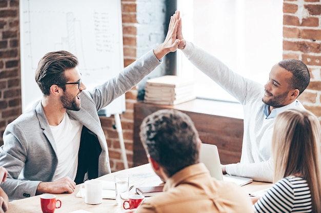 Conseguimos! dois rapazes dando high-five enquanto seus colegas de trabalho estão sentados à mesa do escritório