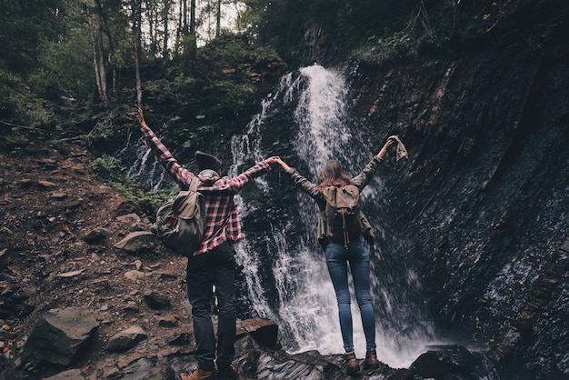 Conseguimos! Retrovisor de corpo inteiro de um jovem casal levantando as mãos em pé perto da cachoeira