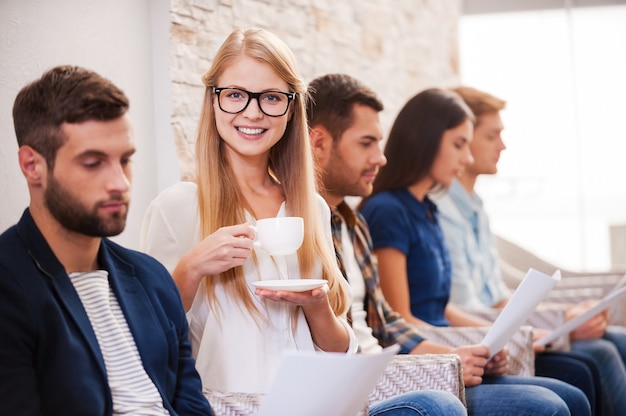 ¡Conseguiré este trabajo! Grupo de jóvenes en ropa casual elegante sentados en una fila en las sillas y sosteniendo papeles mientras hermosa mujer tomando café y sonriendo