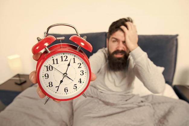 Consejos para convertirse en un madrugador Hombre barbudo hipster cara somnolienta en la cama con reloj despertador Problema con el despertar temprano en la mañana Levantarse con reloj despertador Se quedó dormido otra vez Consejos para despertarse temprano