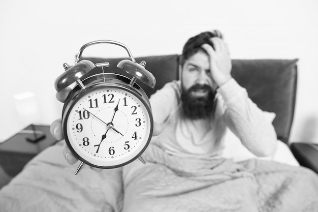 Consejos para convertirse en un madrugador Hombre barbudo hipster cara somnolienta en la cama con reloj despertador Problema con el despertar temprano en la mañana Levantarse con reloj despertador Se quedó dormido otra vez Consejos para despertarse temprano
