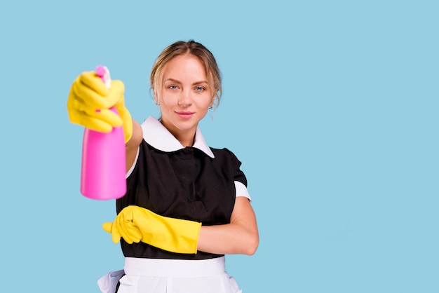 Foto conserje mujer sonriente mostrando botella de spray rosa mirando a la cámara de pie contra la pared azul