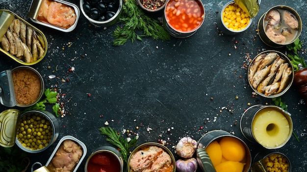 Conservas de legumes feijão peixe e frutas em latas em fundo de pedra preta Estoques de alimentos