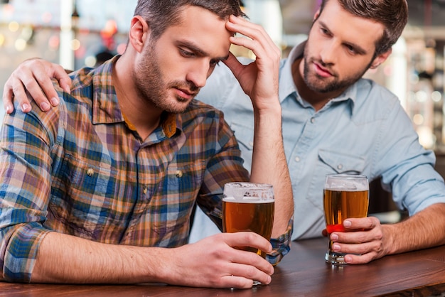 Consolando a su amigo deprimido. Joven deprimido sentado en la barra del bar y sosteniendo la cabeza en la mano mientras es consolado por su amigo sentado cerca de él.