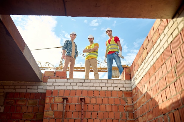 Construção. Vista inferior do engenheiro e dois trabalhadores com capacetes de proteção em um prédio de tijolos em construção, olhando com confiança para a câmera em um dia ensolarado