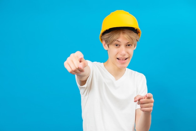 Un constructor adolescente feliz y apuesto con un casco amarillo está mirando a la cámara y te señala con los dedos de ambas manos parado en un estudio con un fondo azul