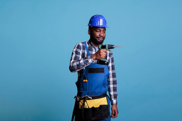 Constructor afroamericano en casco protector uniforme de trabajo con taladro eléctrico sobre fondo azul. Herramienta de trabajo de sujeción profesional de la construcción usando cinturón con martillos.