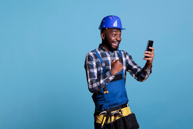 Constructor afroamericano feliz por la noticia del trabajo de renovación recibido por mensaje de teléfono celular. Contratista en uniforme emocionado usando dispositivo móvil en estudio con fondo azul.