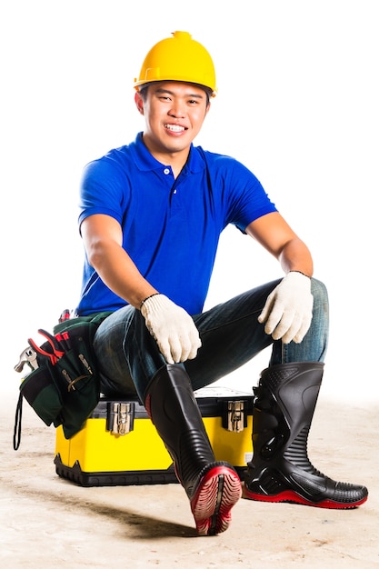 Constructor asiático indonesio o trabajador de la construcción con casco y cinturón de herramientas sentado en la caja de herramientas