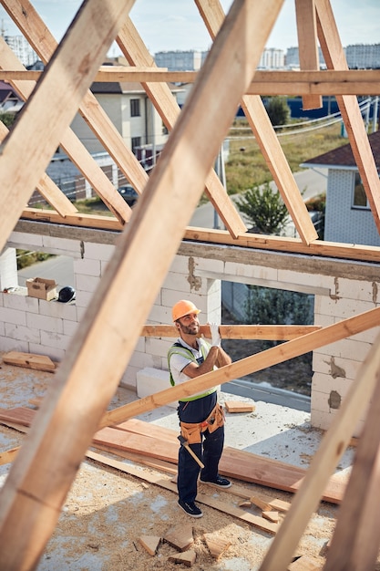 Constructor de aspecto positivo sosteniendo una pieza larga de madera de construcción mientras hace una carcasa de techo