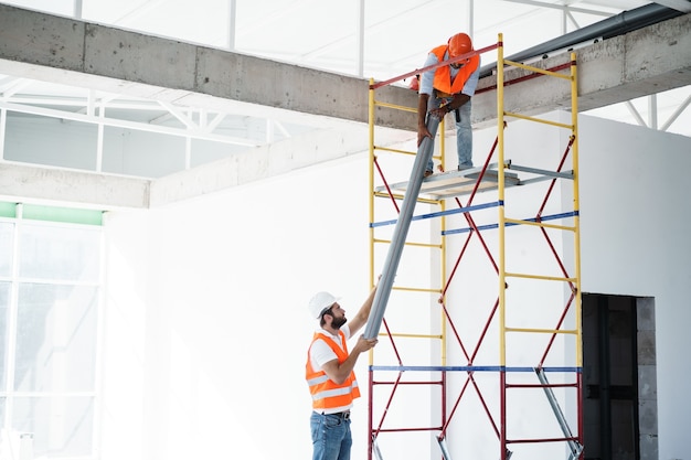 Constructor de dos hombres en uniforme arrastra el tubo en andamios en el sitio de construcción