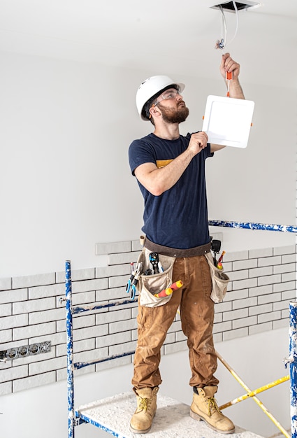 Constructor de electricista con barba trabajador en un casco blanco en el trabajo, instalación de lámparas en altura.