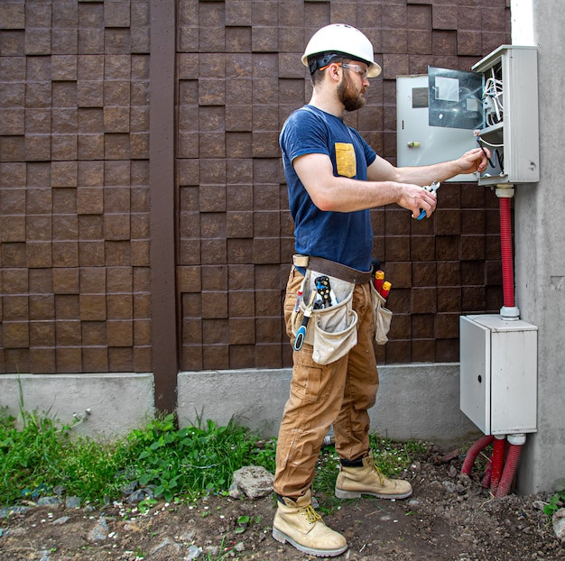 Constructor electricista en el trabajo, examina la conexión del cable en la línea eléctrica en el fuselaje de un cuadro de distribución industrial. Profesional en monos con herramienta de electricista.