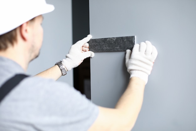 El constructor con guantes y casco elige el color de las baldosas para la pared del apartamento. El hombre aplica una muestra de material de construcción a la pared