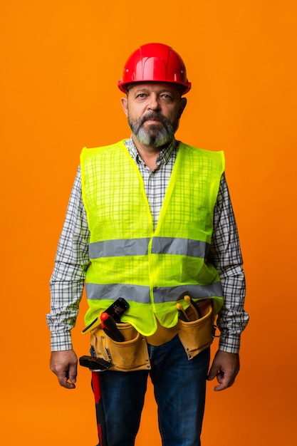 Constructor de hombre barbudo de mediana edad en uniforme y casco con herramientas contra el fondo naranja