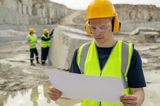 Constructor joven contemporáneo en ropa de trabajo protectora mirando boceto