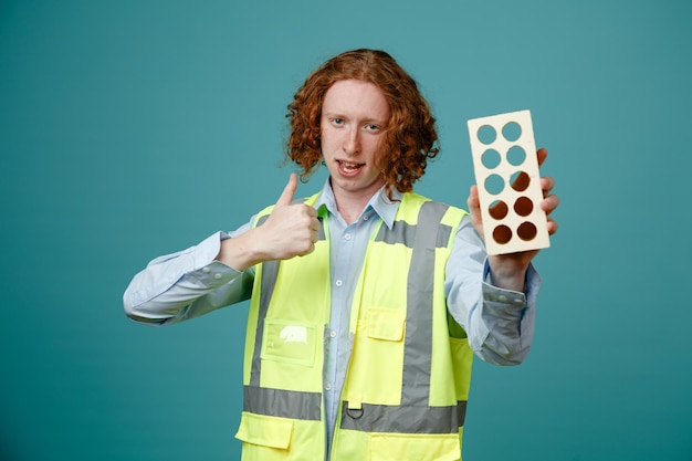 Constructor joven en uniforme de construcción sosteniendo enérgicamente mostrando el pulgar hacia arriba feliz y positivo mirando a la cámara de pie sobre fondo azul.