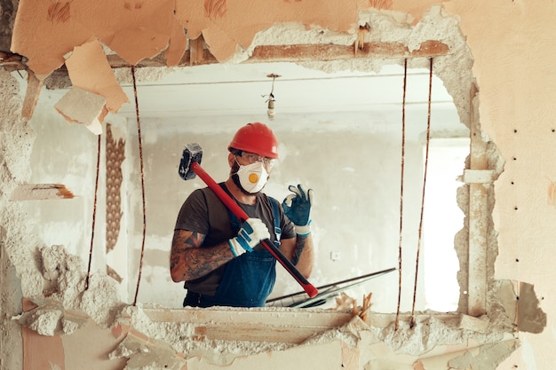 Foto constructor con un martillo en sus manos rompe la pared de cemento