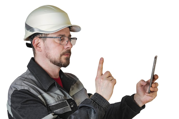 Constructor masculino barbudo amigable positivo en monos casco de seguridad y gafas mira el teléfono y muestra el gesto de la mano Fondo aislado