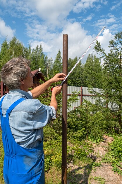 Constructor masculino con una cinta métrica en un sitio de construcción real en la naturaleza. Mide la distancia entre los postes con una cinta métrica.