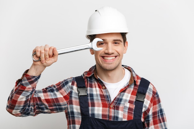 Constructor masculino feliz en casco protector divirtiéndose con llave mientras sobre pared gris