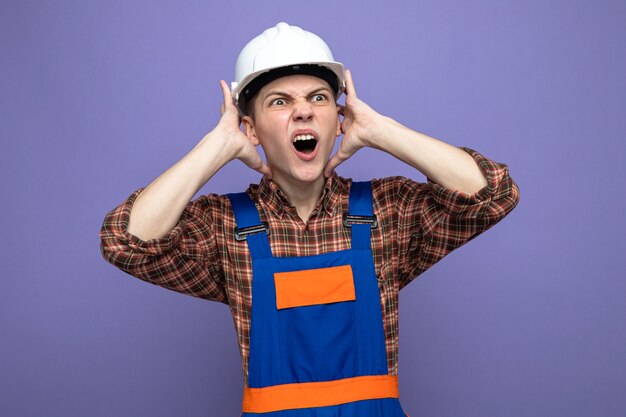 Constructor masculino joven vistiendo uniforme aislado en la pared púrpura