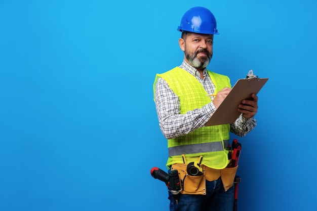 Constructor masculino o manitas en uniforme sosteniendo el portapapeles contra el fondo azul.