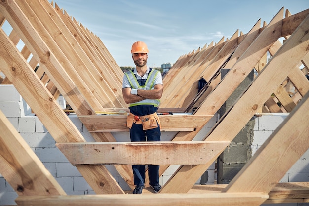 Constructor masculino que parece contento y con uniforme completo y equipo mientras tiene los brazos cruzados sobre el pecho