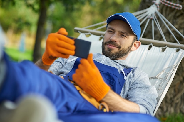 Constructor masculino vistiendo uniforme, tumbado en una hamaca al aire libre y mirando algo con el teléfono inteligente