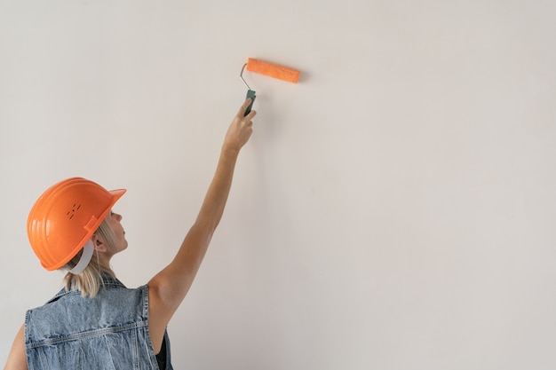 Constructor de mujer hermosa joven en un casco protector naranja en la cabeza. Niña embarazada. Pinta la pared con un rodillo de pintura. Primer plano de una mano y una herramienta. Lugar para el texto