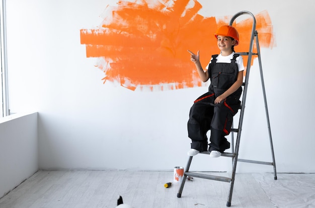 Constructor de niña vestida con ropa de constructor y un casco naranja en la cabeza. sentado en las escaleras señala con el dedo un lugar vacío. Concepto de renovación del hogar.