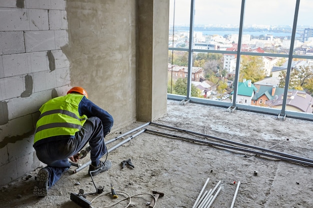 Constructor en ropa de trabajo y casco de seguridad está instalando  tuberías de plástico en el piso del edificio en construcción con ventanas  panorámicas en casa de gran altura | Foto Premium