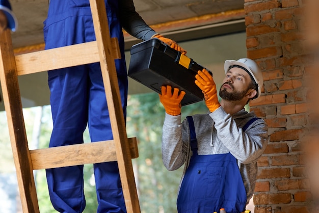 Constructor de sexo masculino joven atractivo en monos y casco dando caja de herramientas a su colega en la escalera