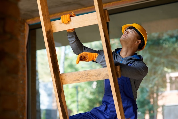 Constructor de sexo masculino joven en monos y casco mirando enfocado, subiendo la escalera