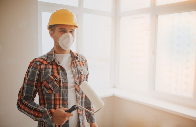 El constructor trabaja en el sitio de construcción y mide el techo. Un trabajador en un casco naranja y un rodillo de pintura pinta la pared.