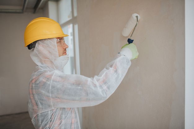 El constructor trabaja en el sitio de construcción. Trabajador con un rodillo de pintura. Lleva un traje protector y una máscara para una cara.