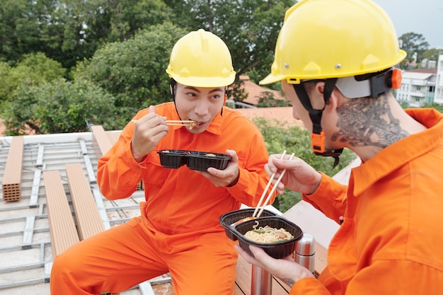 Constructores comiendo sopa de fideos en el sitio de construcción para el almuerzo e intercambiando chistes divertidos